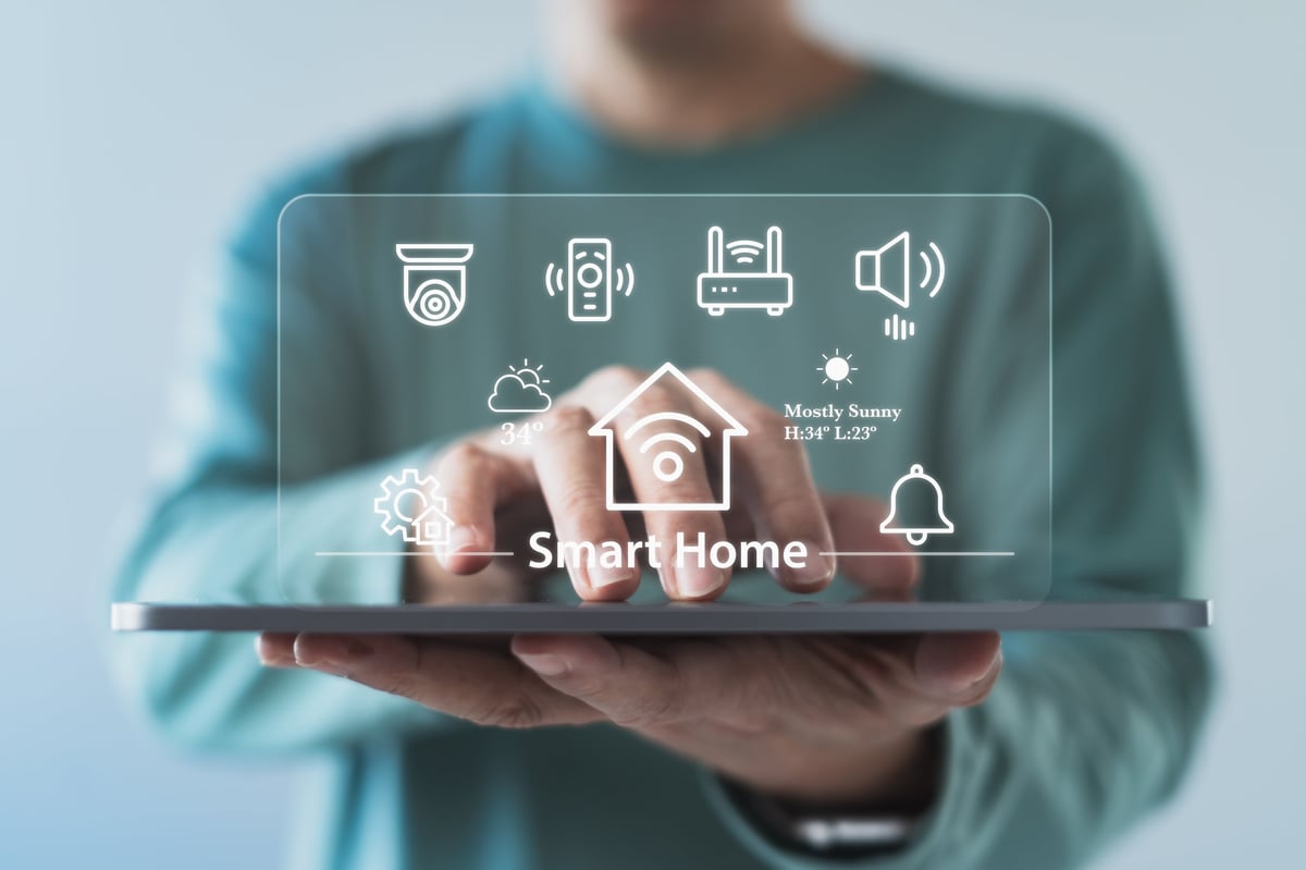 Person using a smart home system interface on a tablet with connected device icons, weather information, security and automation controls for IoT technology and digital home management.